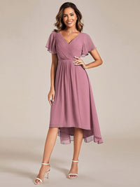 Model in oudroze chiffon jurk met V-hals, fladdermouwen en asymmetrische zoom, met witte sandalen en hand op de heup. 58