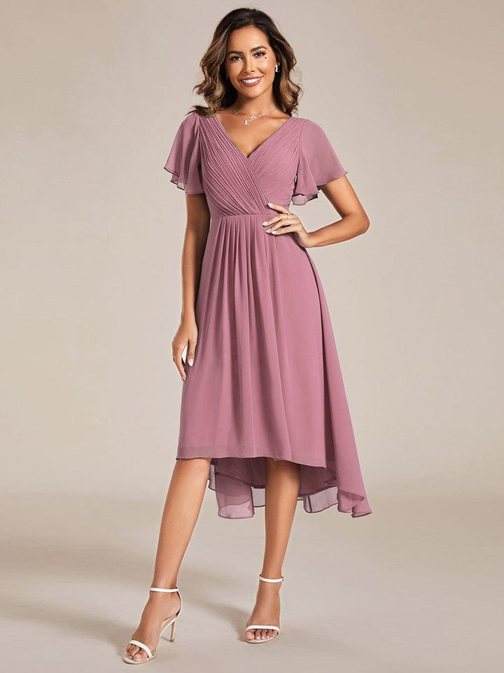 Model in oudroze chiffon jurk met V-hals, fladdermouwen en asymmetrische zoom, met witte sandalen en hand op de heup. 58