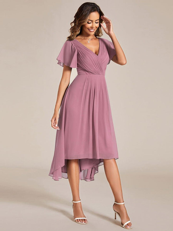 Model in oudroze jurk met asymmetrische zoom, hand bij het hoofd, gecombineerd met witte sandalen met enkelbandje. 62