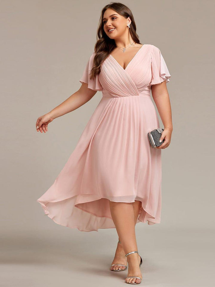 Roze jurk in grotere maat met V-hals en hoog-laag zoom, model draagt zilverkleurige sandalen en een clutch. 31