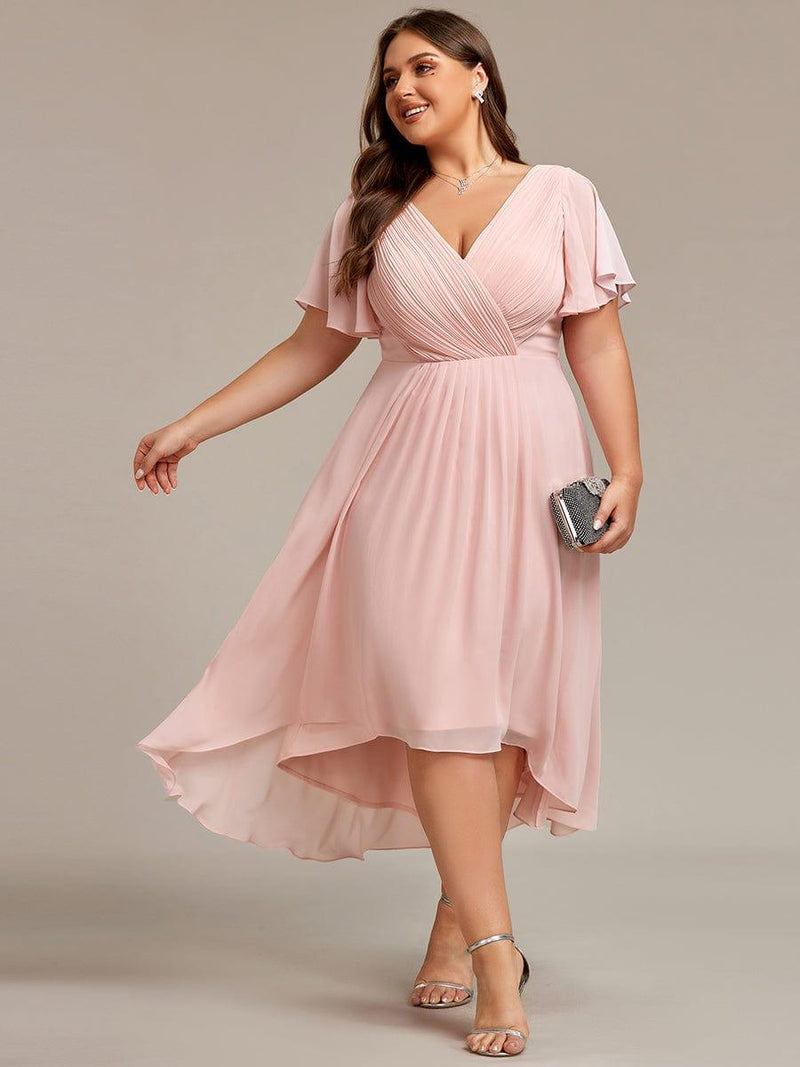 Roze jurk in grotere maat met V-hals en hoog-laag zoom, model draagt zilverkleurige sandalen en een clutch. 31