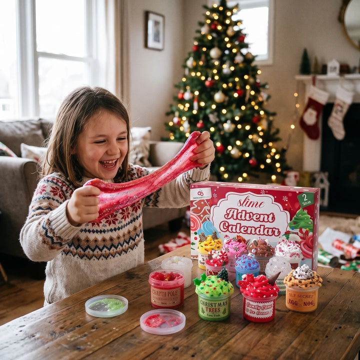 Lachend meisje in kersttrui rekt rode glitterslijm uit naast de doos en geopende potjes, bij de kerstboom 1