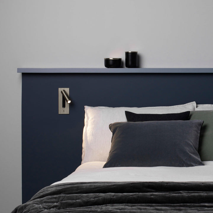 Kleine wandlamp naast het hoofdeinde van een bed met donkerblauwe wand en grijze kussens 30