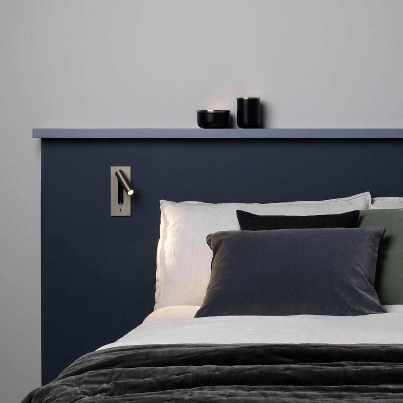 Kleine wandlamp naast het hoofdeinde van een bed met donkerblauwe wand en grijze kussens 30