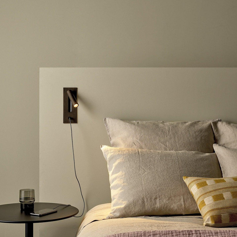 Bronskleurige wandlamp boven een bed, met kabel naar een telefoon op een zwart bijzettafeltje 36
