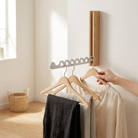 Lichthouten wandkapstok met zilverkleurige arm waaraan drie houten hangers met kleding hangen, terwijl een hand een hanger vastpakt. 3