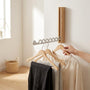 Lichthouten wandkapstok met zilverkleurige arm waaraan drie houten hangers met kleding hangen, terwijl een hand een hanger vastpakt. 3