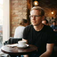 Man met de bril met doorzichtig grijs montuur zit aan een rond tafeltje in een café bij een kop koffie. 4