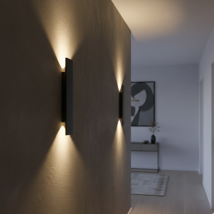 Zwarte wandlampen op een gang binnenshuis, met warm licht boven en onder tegen een gestuukte muur en een kunstwerk op de achtergrond. 3