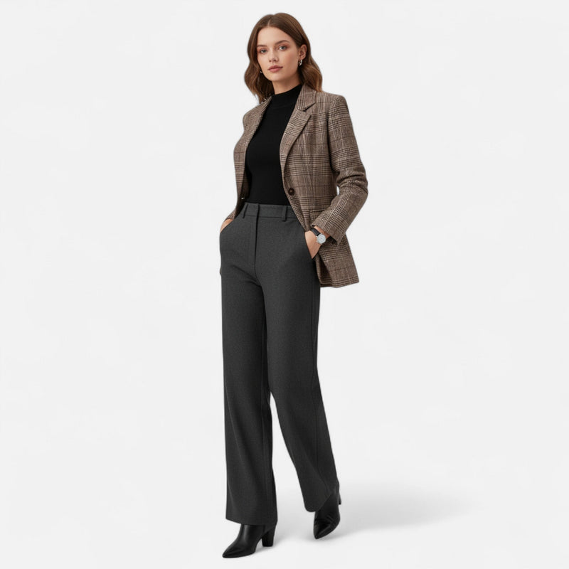 Model in volledige outfit met de blazer over een zwarte top, grijze broek met wijde pijpen en zwarte laarzen, handen in de zakken. 2