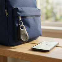 Druppelvormige tracker aan de rits van een blauwe rugzak, naast een telefoon met kaart op houten tafel 1