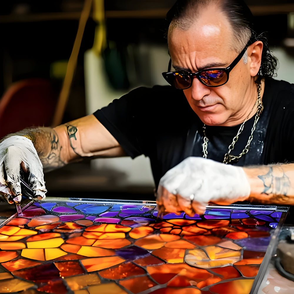Een ambachtsman met zonnebril en handschoenen legt gekleurde glasstukjes in een mozaïek aan een werktafel 5