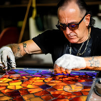 Een ambachtsman met zonnebril en handschoenen legt gekleurde glasstukjes in een mozaïek aan een werktafel 5
