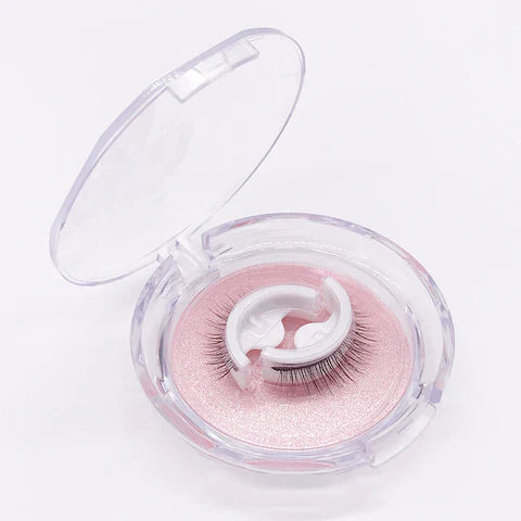 Open rond doosje met doorzichtig deksel, met een paar wimpers op een wit houdertje tegen een roze glitterbodem. 1