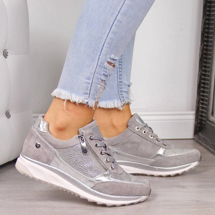 Grijze sneakers met zilverkleurige accenten en zijrits, gedragen door een model met gerafelde spijkerbroek naast een gecapitonneerd wit kussen. 2