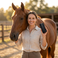 Lachende vrouw in linnen blouse houdt de zwarte borstel omhoog naast het hoofd van een kastanjebruin paard in de wei. 2