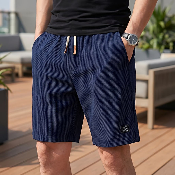 Man in donkerblauwe wafelshort met wit trekkoord en leren uiteinden, hand in de zak, staand op een houten terras. 7