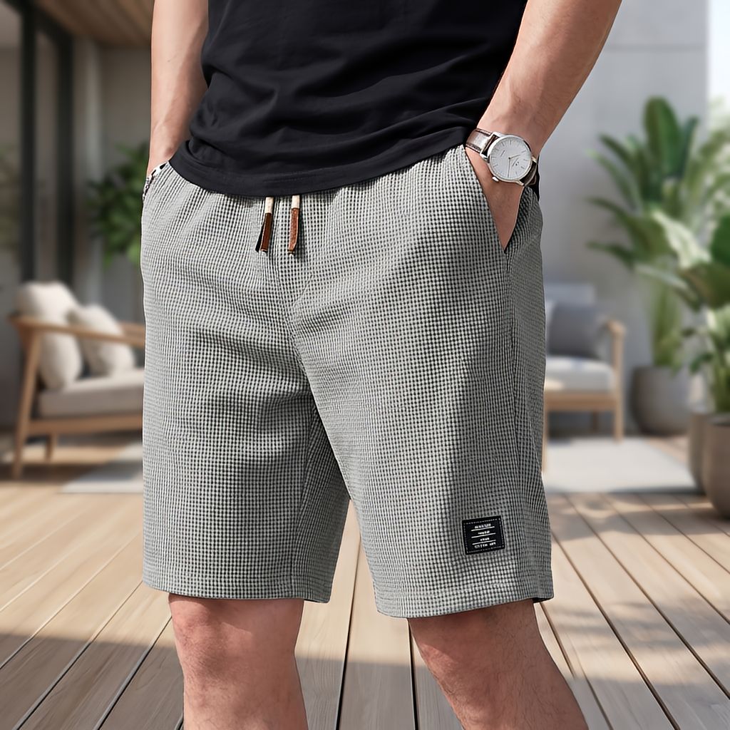 Man draagt een grijze wafelshort met trekkoord en horloge, hand in de zak, op een terras met planten en loungestoel. 6