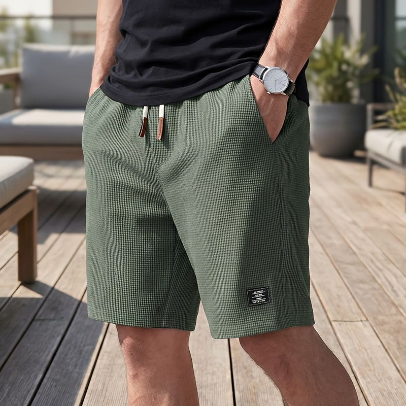 Man in groene wafelshort met wit trekkoord met bruine uiteinden, hand in de zak, staand op een houten terras. 5
