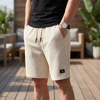 Man in beige wafelshort met crèmekleurig trekkoord en bruine uiteinden, een hand in de zak, op een houten terras. 8