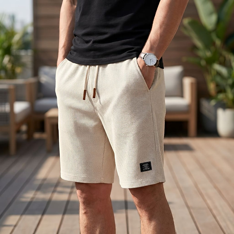 Man in beige wafelshort met crèmekleurig trekkoord en bruine uiteinden, een hand in de zak, op een houten terras. 8