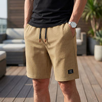 Man in zandkleurige wafelshort met zwart trekkoord met bruine uiteinden, hand in de zak, op een zonnig terras. 4