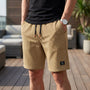 Man in zandkleurige wafelshort met zwart trekkoord met bruine uiteinden, hand in de zak, op een zonnig terras. 4
