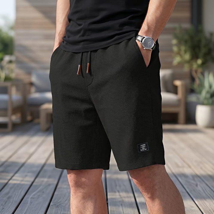 Man draagt een zwarte wafelshort met trekkoord en horloge, een hand in de zak, staand op een houten terras. 3