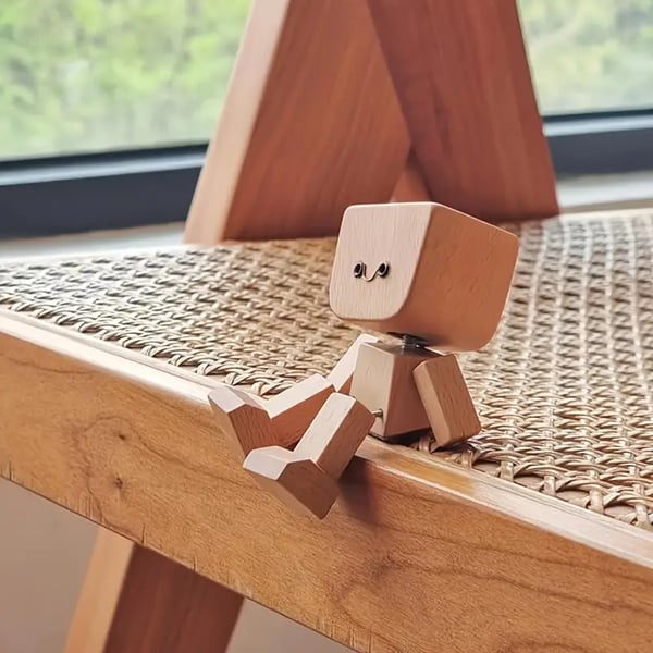 Klein houten figuurtje met lachend gezichtje zit op de rand van een rotan stoelzitting naast een raam. 5