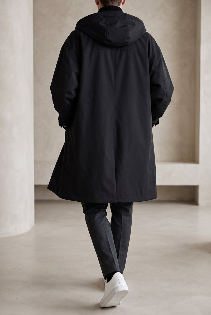 Achterkant van de zwarte lange jas met capuchon, gedragen door een model met grijze broek en witte sneakers 7