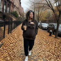 Vrouw in donkerbruine sherpahoodie met capuchon op loopt over een met herfstbladeren bedekte straat met een baby in het paneel. 5