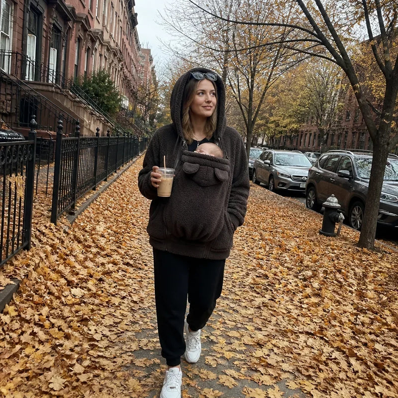 Vrouw in donkerbruine sherpahoodie met capuchon op loopt over een met herfstbladeren bedekte straat met een baby in het paneel. 5