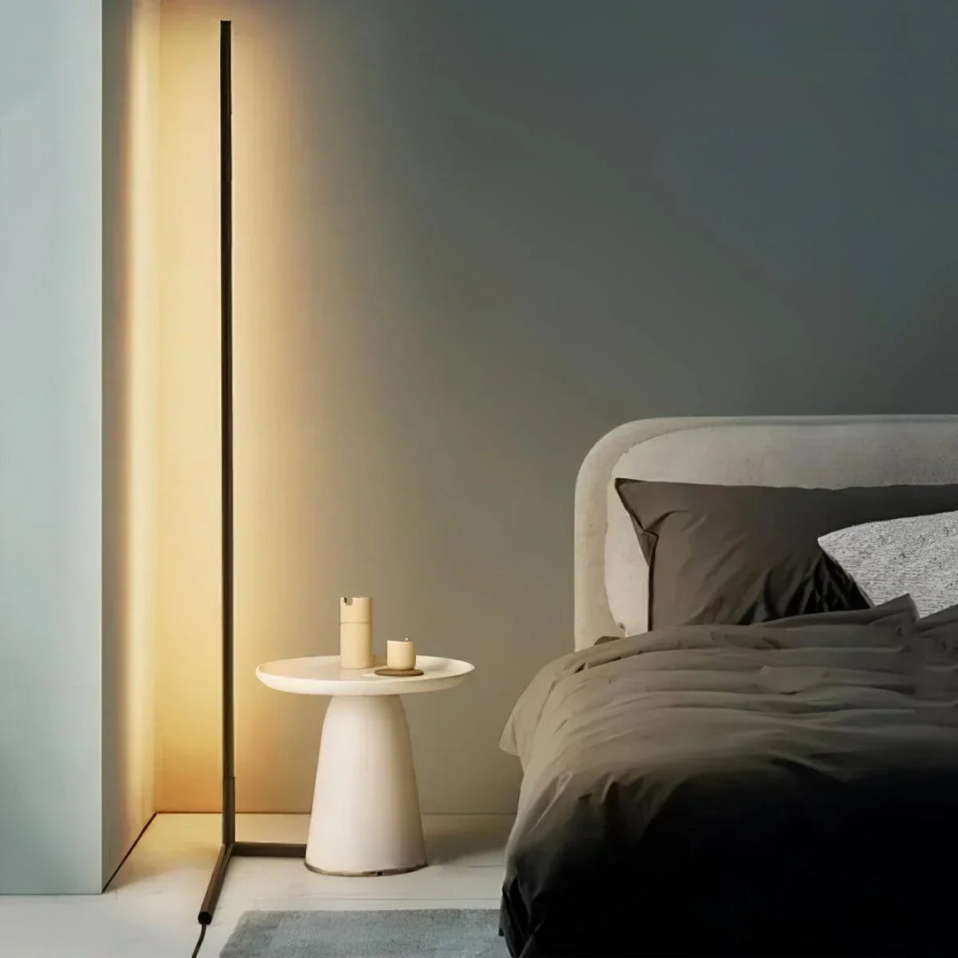 Dunne zwarte vloerlamp met warm licht naast een ronde witte bijzettafel en een bed met donkere lakens 1
