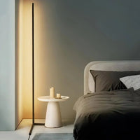 Dunne zwarte vloerlamp met warm licht naast een ronde witte bijzettafel en een bed met donkere lakens 1