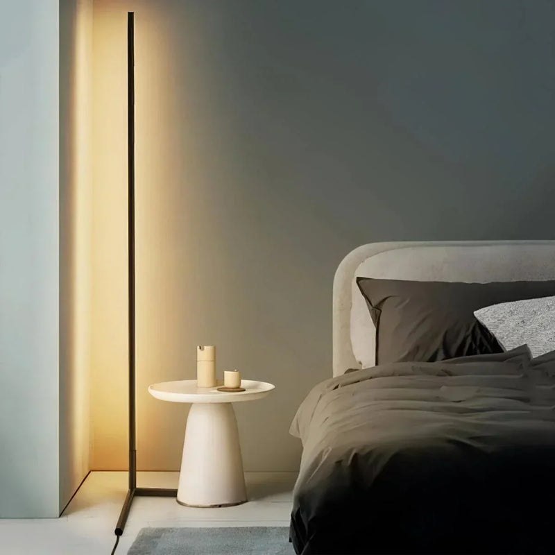 Dunne zwarte vloerlamp met warm licht naast een ronde witte bijzettafel en een bed met donkere lakens 1
