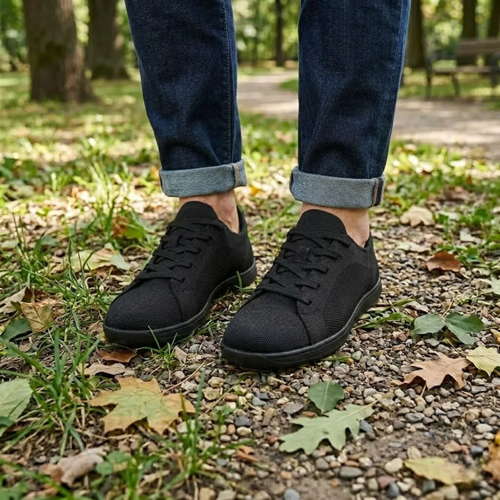 Zwarte gebreide sneakers gedragen met een spijkerbroek met opgerolde pijpen, staand op een bospad met grind en herfstbladeren. 11