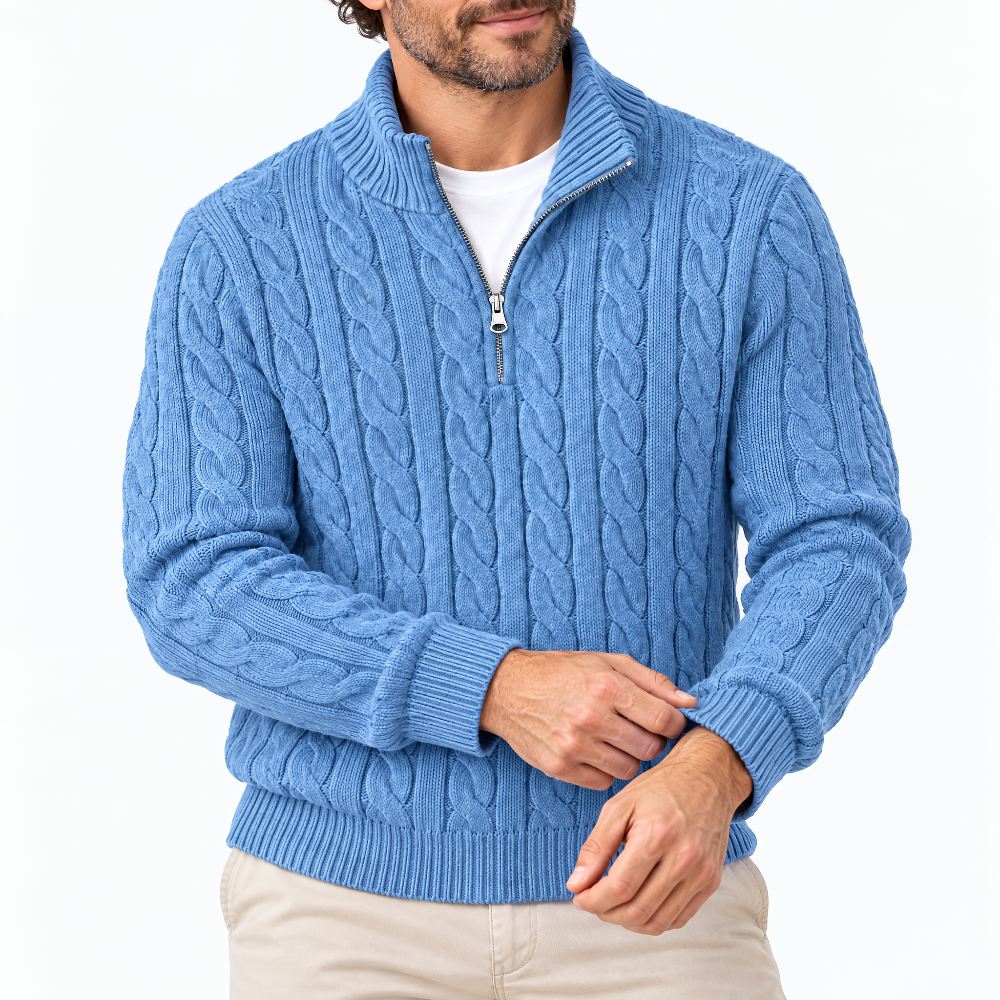 Man in blauwe kabeltrui met halve rits, handen aan de manchet, op witte achtergrond met beige broek. 1