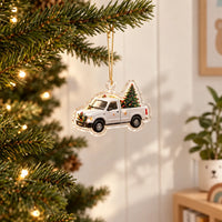 Transparant kerstornament van een witte pick-up met kerstboom in de laadbak, aan een gouden koordje in een verlichte kerstboom gehangen. 1