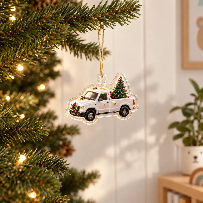 Transparant kerstornament van een witte pick-up met kerstboom in de laadbak, aan een gouden koordje in een verlichte kerstboom gehangen. 1