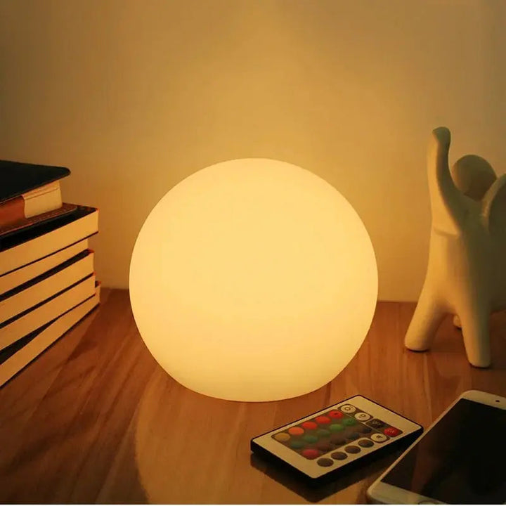 Warm gloeiende bollamp op een houten tafel naast een stapel boeken, een beeldje, een smartphone en de afstandsbediening met gekleurde knoppen. 1