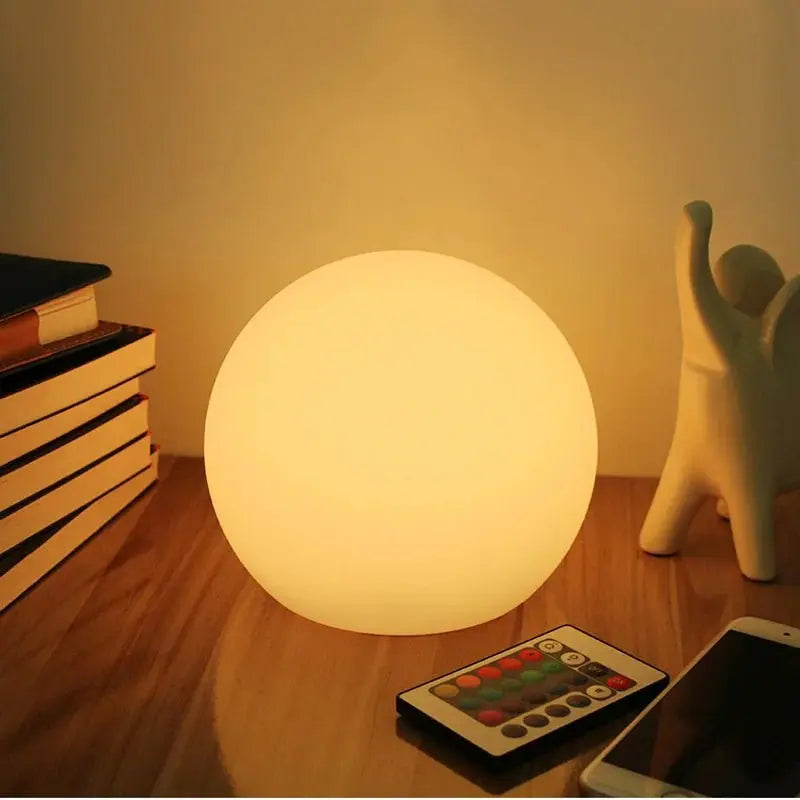 Warm gloeiende bollamp op een houten tafel naast een stapel boeken, een beeldje, een smartphone en de afstandsbediening met gekleurde knoppen. 1