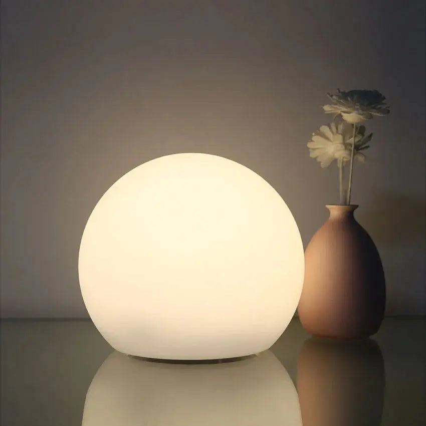 Zacht wit verlichte bollamp op een glanzend oppervlak, naast een bruine vaas met twee bloemen. 2