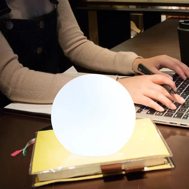 Helder wit oplichtende bollamp op een notitieboek, terwijl iemand op de achtergrond typt op een laptop en een pen vasthoudt. 7