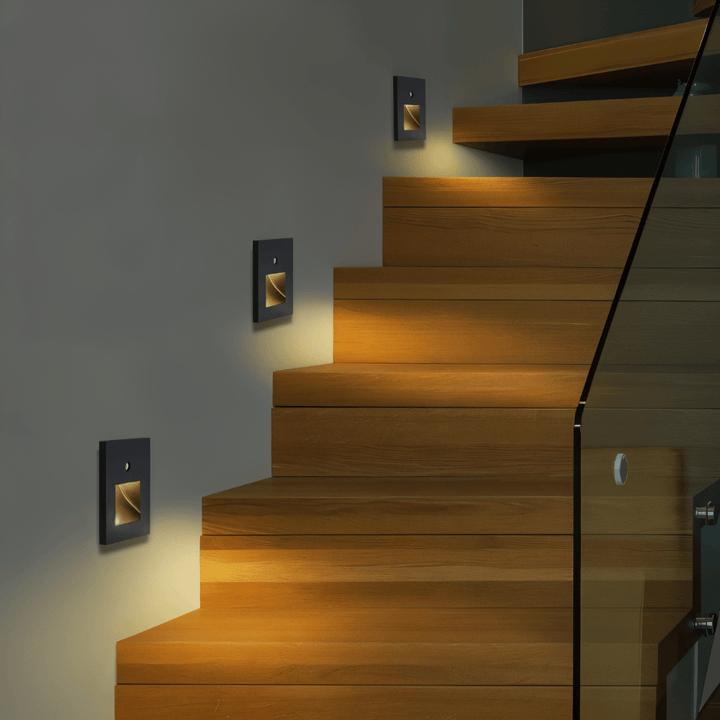 Rij vierkante wandlampjes langs een houten trap met glazen balustrade, die de treden warm verlichten 3