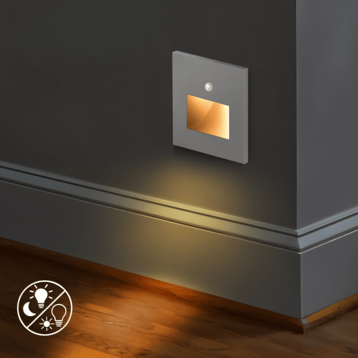 Grijs wandlampje met sensor en gloeiend vakje boven een plint, warm licht op de houten vloer 5