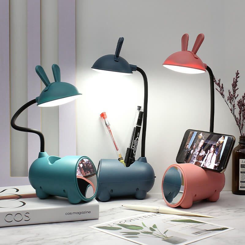 Drie brandende konijnenlampen in groen, donkerblauw en roze op een bureau, met pennen in de houder en een telefoon op de standaard. 1