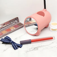 Roze lampvoet met ronde spiegel aan de voorkant, omringd door make-up zoals een oogschaduwpalet, lippenstift en een haarstrikje. 3