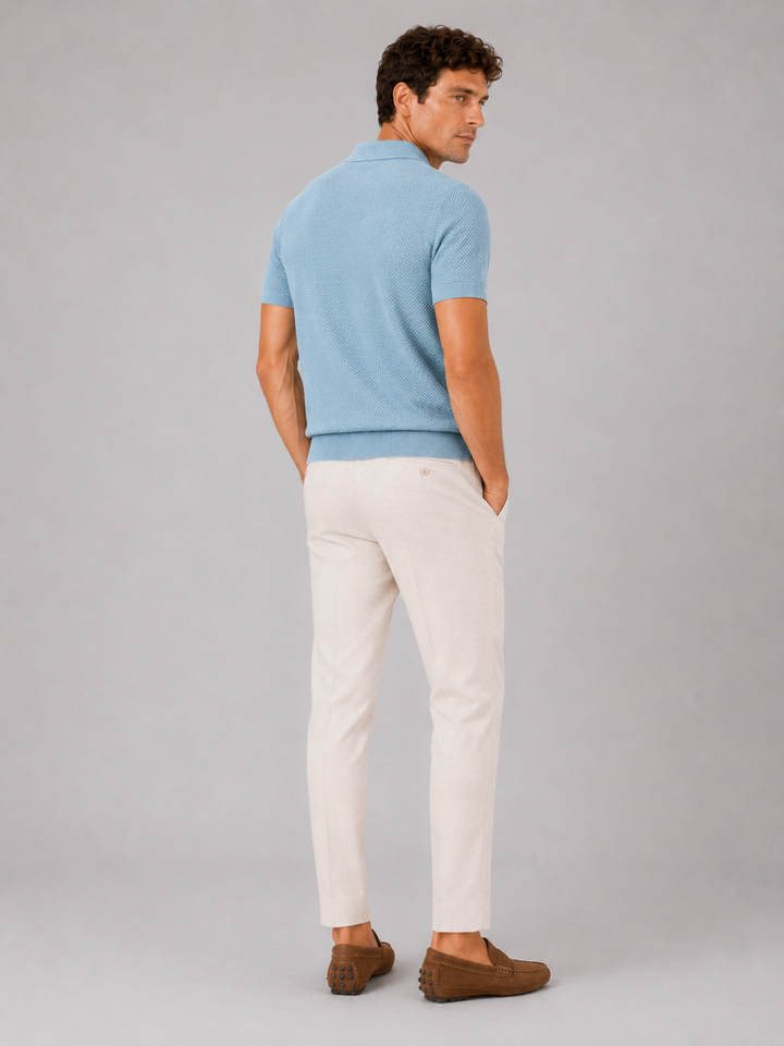 Achterkant van het lichtblauwe poloshirt, model kijkt over zijn schouder, met crèmekleurige broek en bruine loafers 4