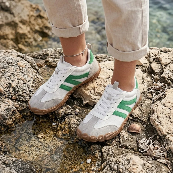 Vrouwenvoeten in witte sneakers met groene strepen, staand op rotsen bij helder water, met opgerolde linnen broek. 29
