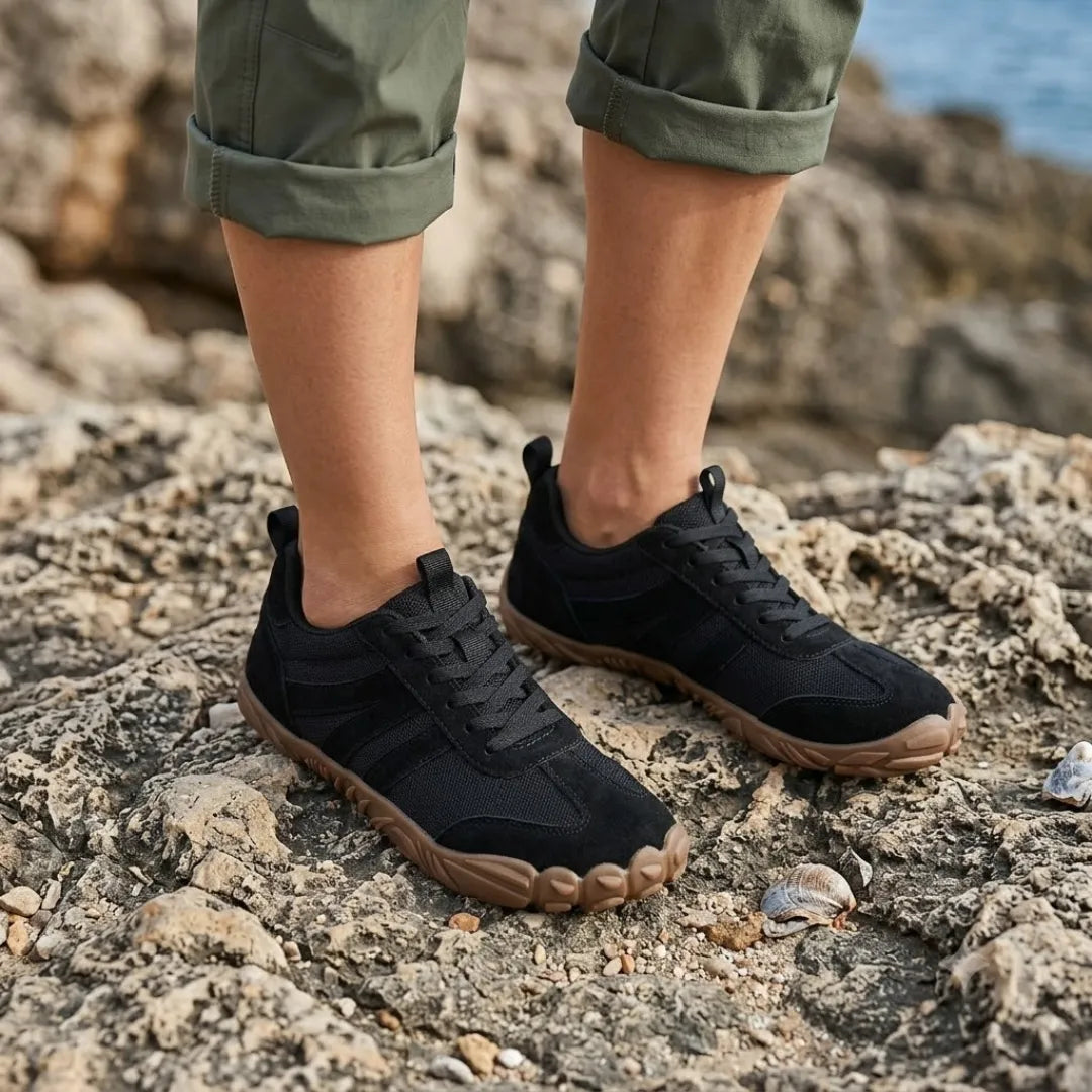Zwarte barefoot schoenen gedragen door een model met opgerolde broekspijpen, staand op ruwe rotsen bij de zee. 20